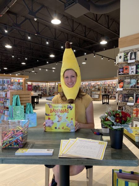 Author Carrie Tillotson wearing a banana hat at a book signing event, holding a copy of the book Counting to Bananas.